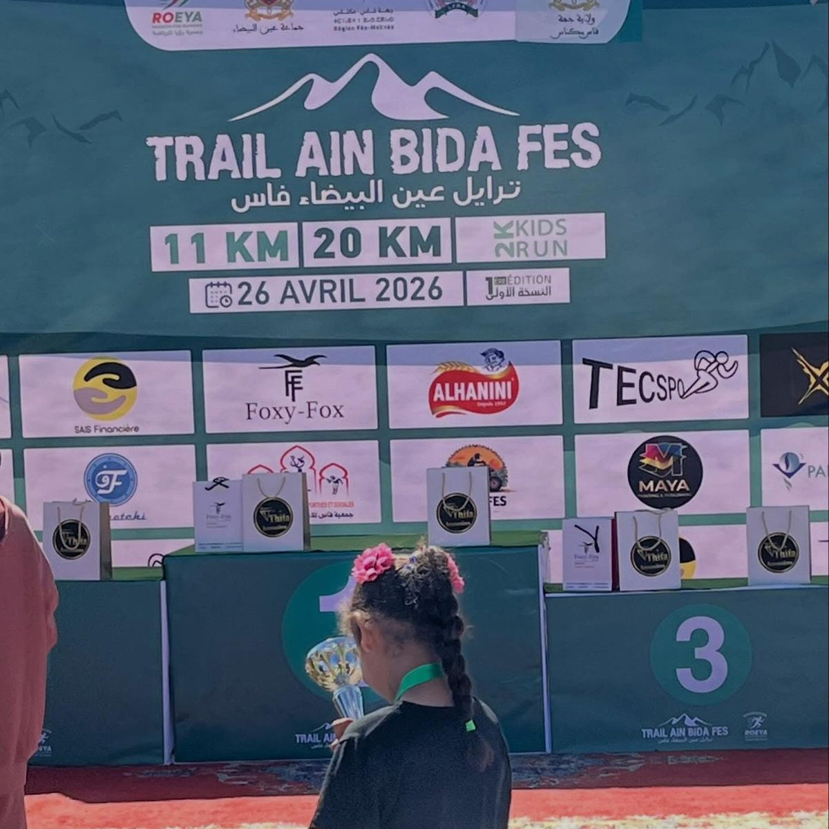 Person standing in front of a large event banner for 'Trail Ain Bida Fes' with various sponsors displayed.